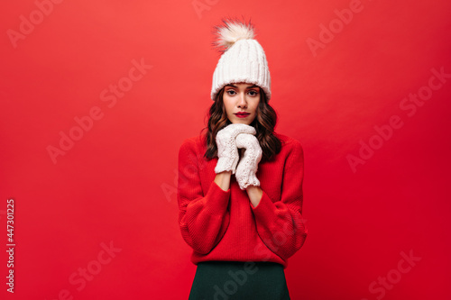 Curly woman in red sweater  knitted hat and mittens looks into camera. Lady in warm winter outfit poses on isolated bright background.