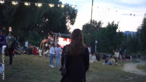Back view of young stylish woman stamding and looking movie in open-air cinema festival alone.