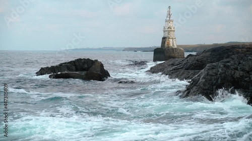 Sea waves hitting and splashing against dark gray cliffs. Old white lighthouse and distant coastline in the background. Cloudy day. Ahtopol on Black sea coast, Bulgaria 