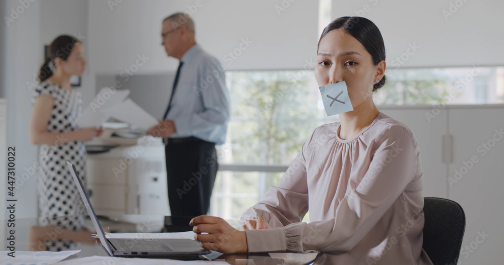 Obraz premium Asian employee working on laptop with drawn cross sticker on her mouth