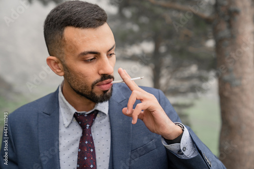 a man with a beard smokes a cigarette standing in the woods by a tree. 