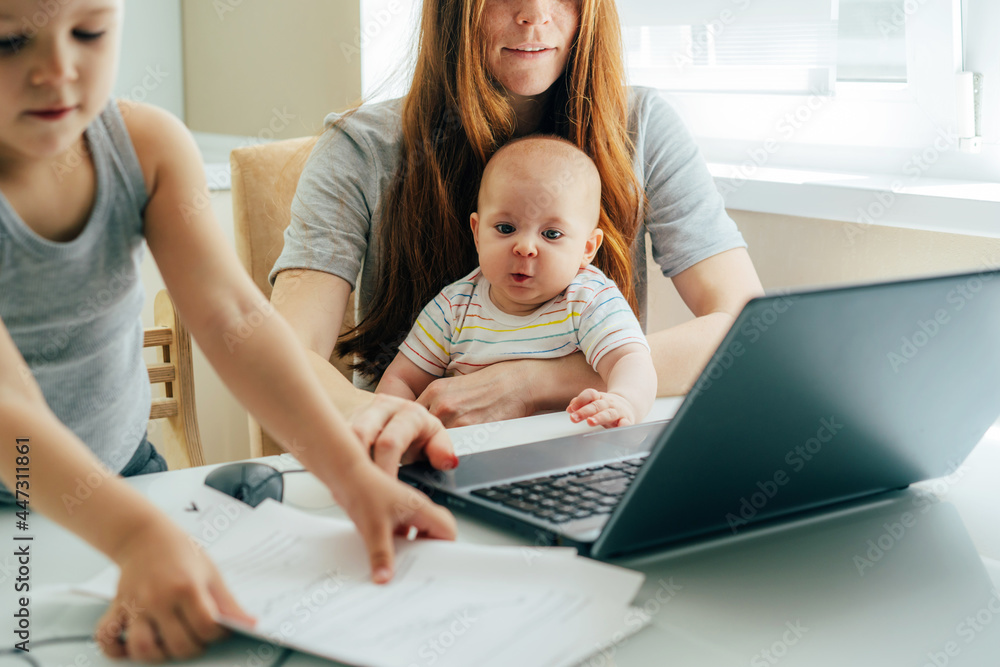 Young focused business mom with two children working remotely on a laptop at home office. Young focused business mom with two children working remotely on a laptop at home office.