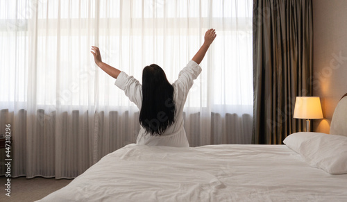 One woman is waking up and stretching her arms in bed.An Asian lady in white top, with bed room and white window curtain background in the early morning with sun light. 