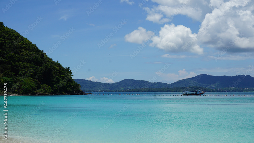 View of the sea in Phuket, Thailand. There are bright blue skies, mountains, and sandy beaches.