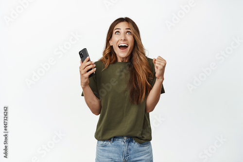 Happy middle aged woman holding mobile phone in hand and celebrating, scream from joy and excitement, looking up and smiling excited, standing against white background
