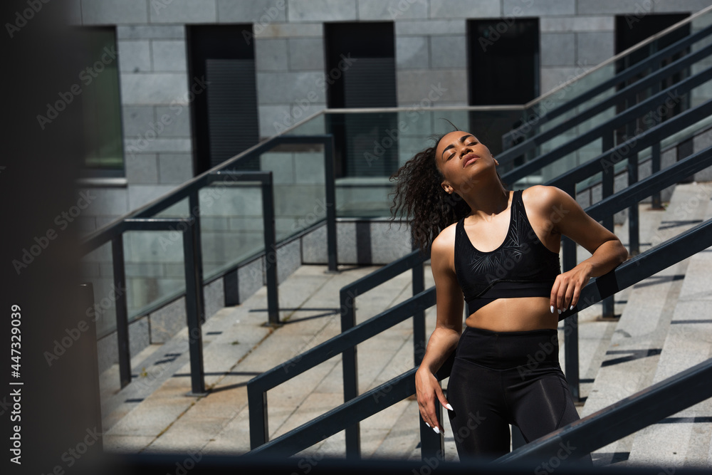 Obraz premium Pretty african american sportswoman with closed eyes standing on stairs near railing
