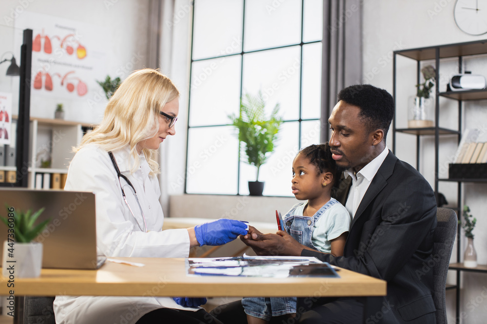 Caucasian doctor checking oxygen saturation of little african girl using modern pulse oximeter. Cute patient sitting on knees af her caring father. Family visiting doctor for controlling health. Caucasian doctor checking oxygen saturation of little african girl using modern pulse oximeter. Cute patient sitting on knees af her caring father. Family visiting doctor for controlling health.