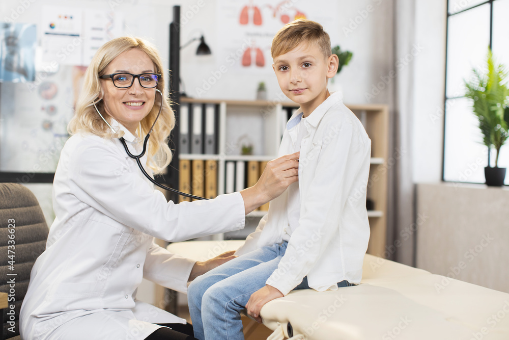 Obraz premium Competent caucasian doctor examining heart rate of teenage boy using stethoscope. Female specialist in white lab coat and eyeglasses caring about little patient.