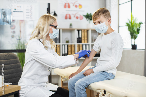 Female doctor in medical mask, gloves and eyeglasses doing vaccination for coronavirus to male patient. Young boy getting immunite at modern clinic