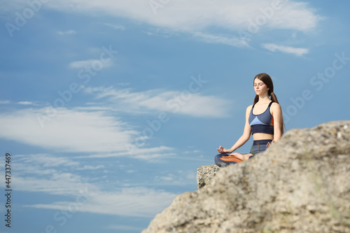 Teenage girl meditating on cliff against blue sky. Space for text