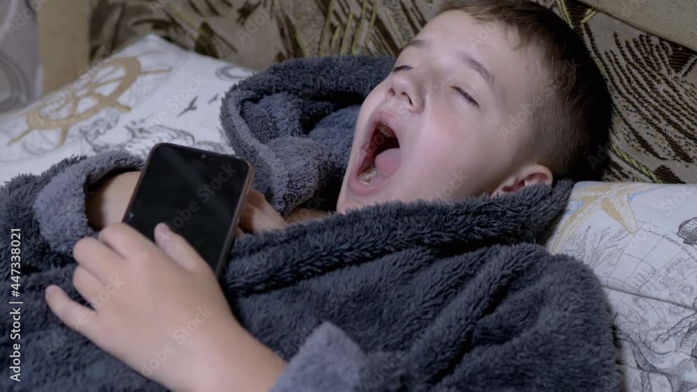 Yawning Child in Home Gray Dressing Gown Falls asleep with Smartphone in Hands. A happy boy holds a mobile phone in his hands. A teenager is dependent on modern gadgets and tablets. 4K. Slow motion. Yawning Child in Home Gray Dressing Gown Falls asleep with Smartphone in Hands. A happy boy holds a mobile phone in his hands. A teenager is dependent on modern gadgets and tablets. 4K. Slow motion.