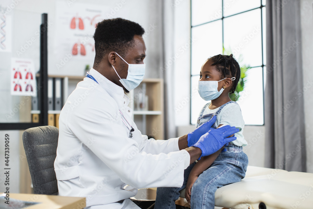 Fototapeta premium Competent male pediatrician in medical mask and gloves making Covid-19 vaccination in shoulder of little african girl. Concept of immunisation, protection and medicine.