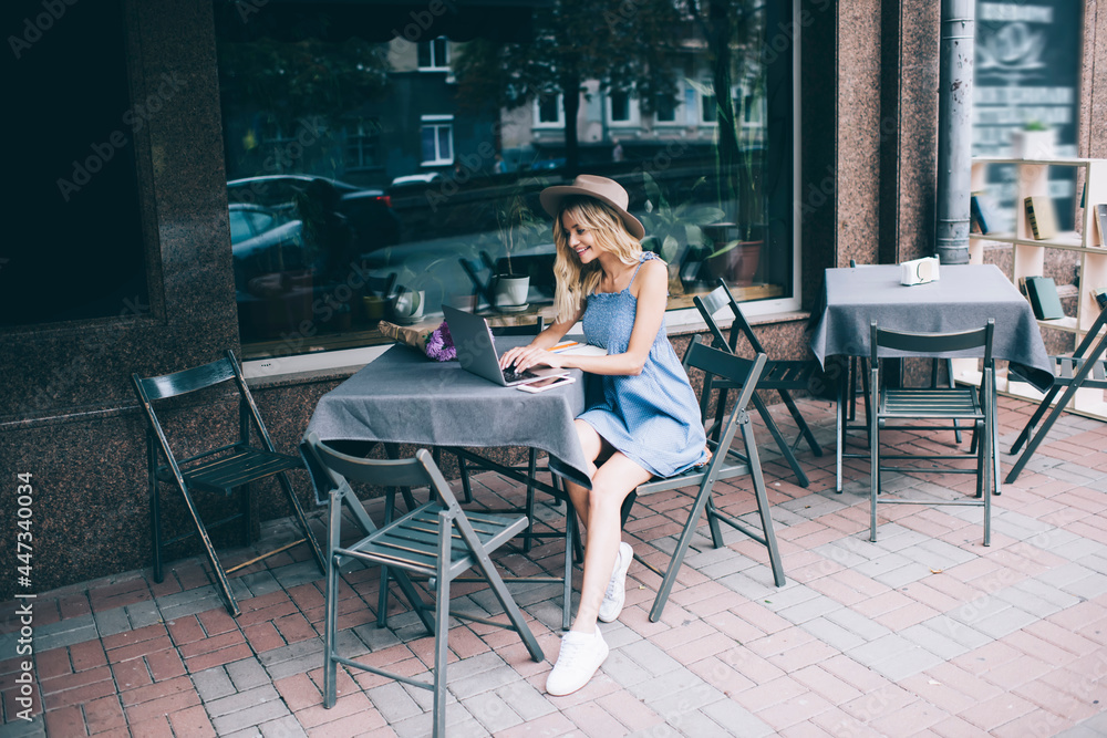 Pretty Caucasian woman in stylish hat browsing website for chatting in media networks via laptop application, smiling hipster girl searching funny content publication via app on digital netbook