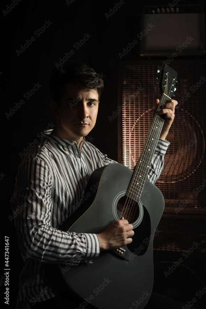 Obraz premium Photo of man sitting and playing acoustic guitar in recording studio on dark background with light effect. Repetition of the rock music of the guitarist. Rehearsal base