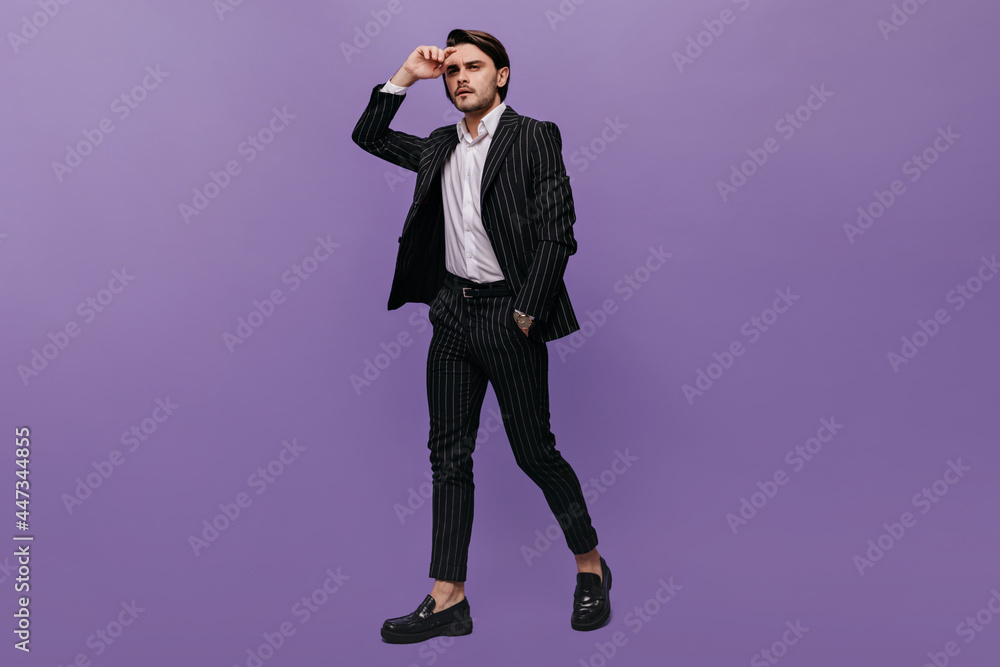 Handsome young gentleman with brunette hair, stylish classic suit and black shoes looking away while posing isolated on violet background Handsome young gentleman with brunette hair, stylish classic suit and black shoes looking away while posing isolated on violet background