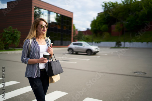 Caucasian hipster girl with shopping bags and caffeine beverage walking at urban street, millennial female fashion blogger holsing modern cellphone technology for networking and takeaway coffee to go © BullRun