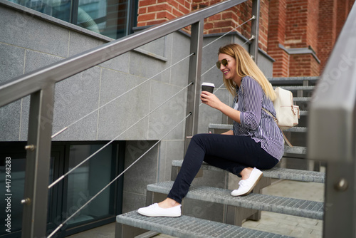 Charming hipster girl with takeaway caffeine beverage in hand resting at city stairs and laughing during break, cheerful female tourist with coffee to go smiling at urbanity enjoying leisure
