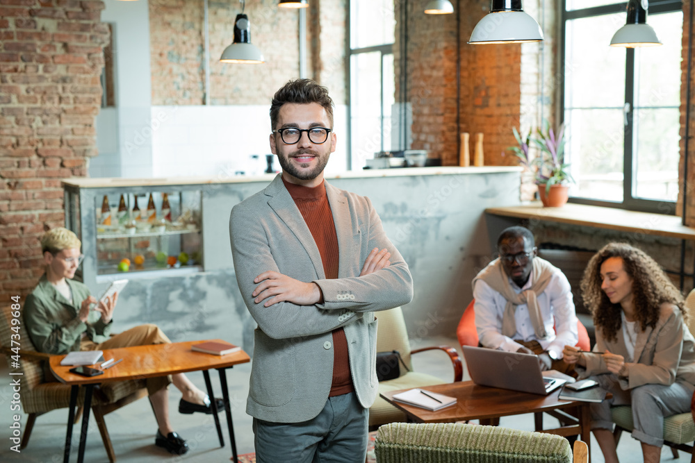 Young smiling cross-armed businessman standing in front of working colleagues Young smiling cross-armed businessman standing in front of working colleagues