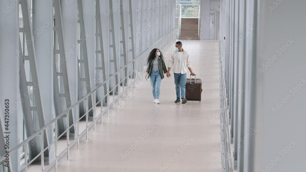Multiracial couple african man and hispanic woman wearing medical masks holding hands walking in airport terminal to board plane journey travel carrying suitcase, afro husband meets wife after arrival Multiracial couple african man and hispanic woman wearing medical masks holding hands walking in airport terminal to board plane journey travel carrying suitcase, afro husband meets wife after arrival