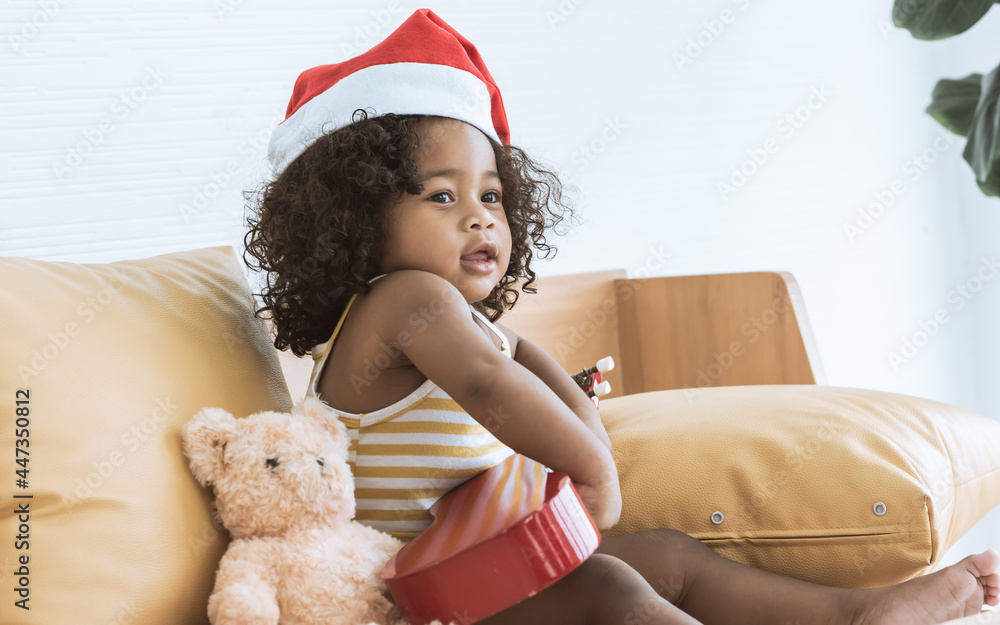 Little cute African black girl wearing Santa hat, sitting on sofa in living room at home, playing ukulele celebrate Christmas or new year party in December with happiness. Kid and Education concept. Little cute African black girl wearing Santa hat, sitting on sofa in living room at home, playing ukulele celebrate Christmas or new year party in December with happiness. Kid and Education concept.