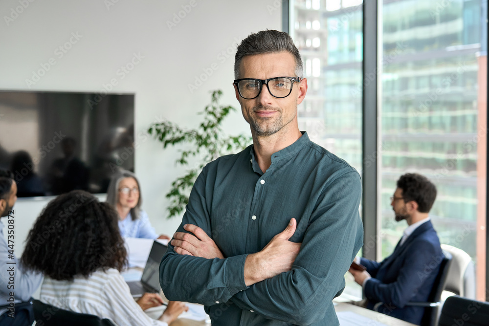 Smiling confident mature businessman leader looking at camera standing in office at team meeting. Male corporate leader ceo executive manager wearing glasses posing for business portrait arms folded. - obrazy, fototapety, plakaty 