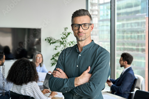 Smiling confident mature businessman leader looking at camera standing in office at team meeting. Male corporate leader ceo executive manager wearing glasses posing for business portrait arms folded. photo
