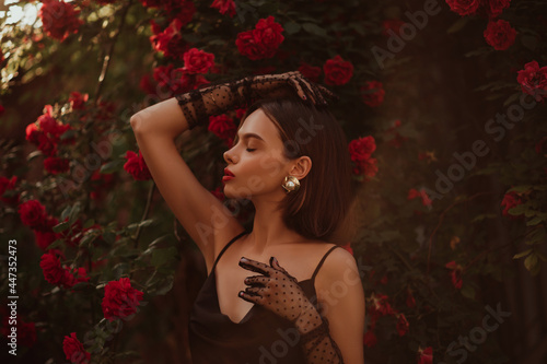 Beautiful fashionable woman with red lips makeup wearing trendy polka dot tulle gloves, vintage pearl earrings, black dress, posing outdoor, in blooming rose garden. Copy, empty space for text