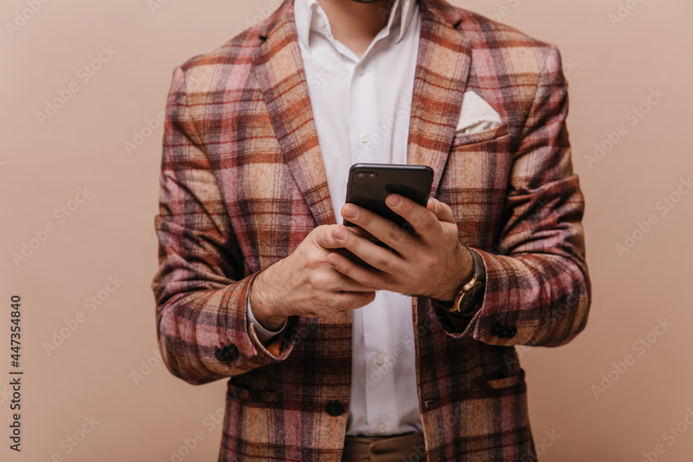 Fototapeta premium Faceless photo of elegant man in white shirt and plaid brown blazer, holding phone and posing isolated on beige background