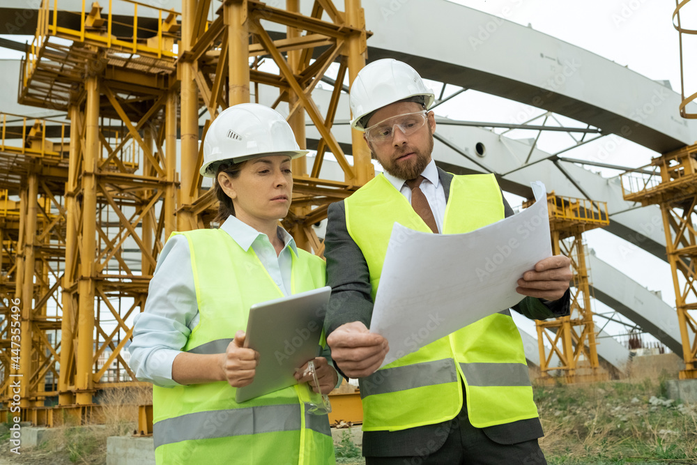 Young architect in work helmet examining blueprint and discussing it with his colleague while she using tablet pc outdoors Young architect in work helmet examining blueprint and discussing it with his colleague while she using tablet pc outdoors