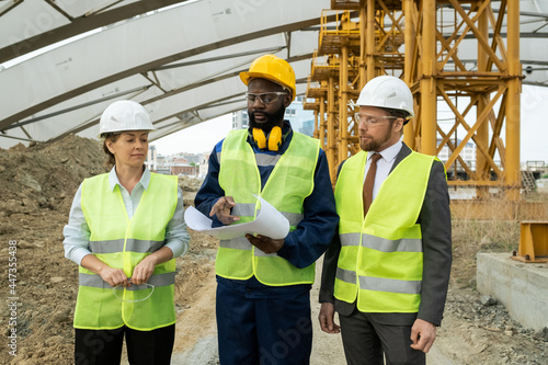 Group of engineers examining blueprint and discussing new construction project in team while walking through construction site © pressmaster