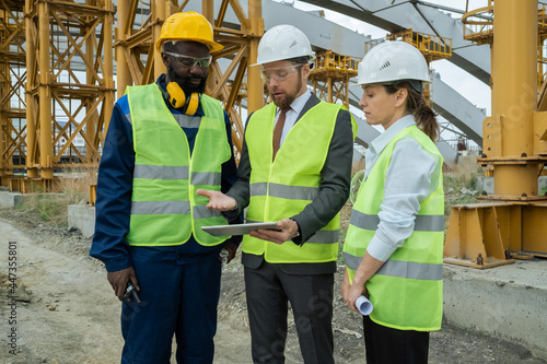 Group of construction workers in work helmets discussing project on digital tablet while standing on construction site