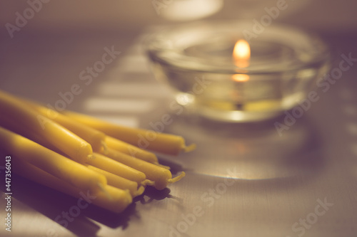 Heap of new yellow candles on the table. One low candle burns in a glass round candlestick in blurred background