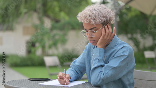 African Woman Thinking while Writing on Paper in Outdoor Cafe