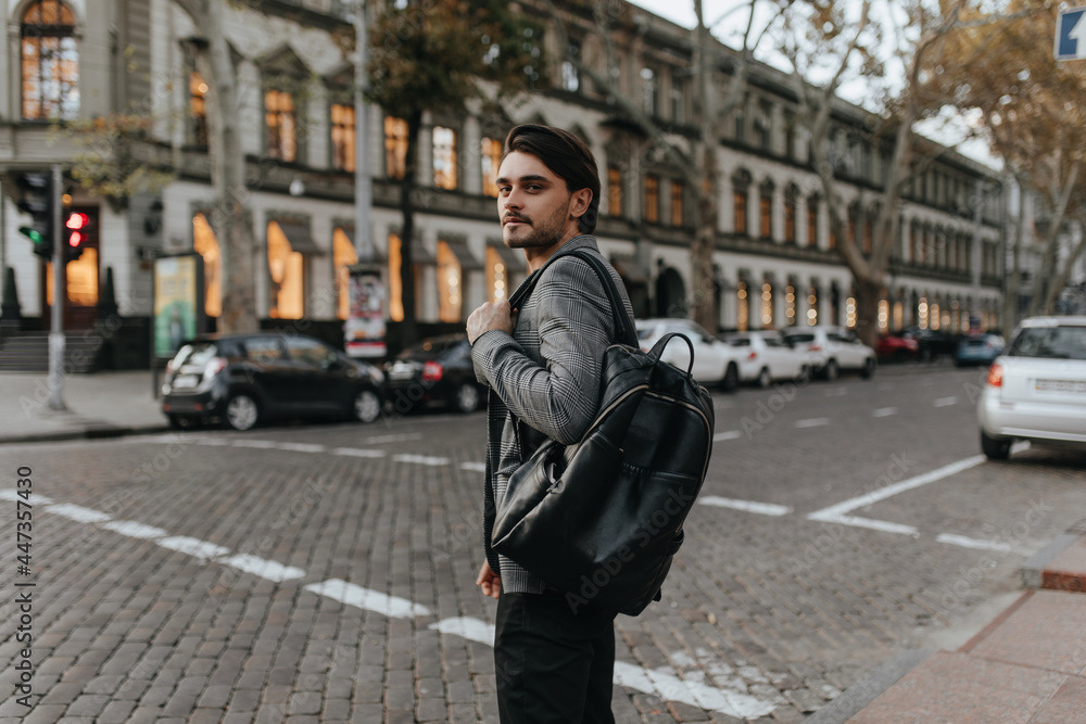 Lifestyle portrait of charming young brunette in grey blazer and black pants, looking into camera and holding backpack. Man posing against day city background