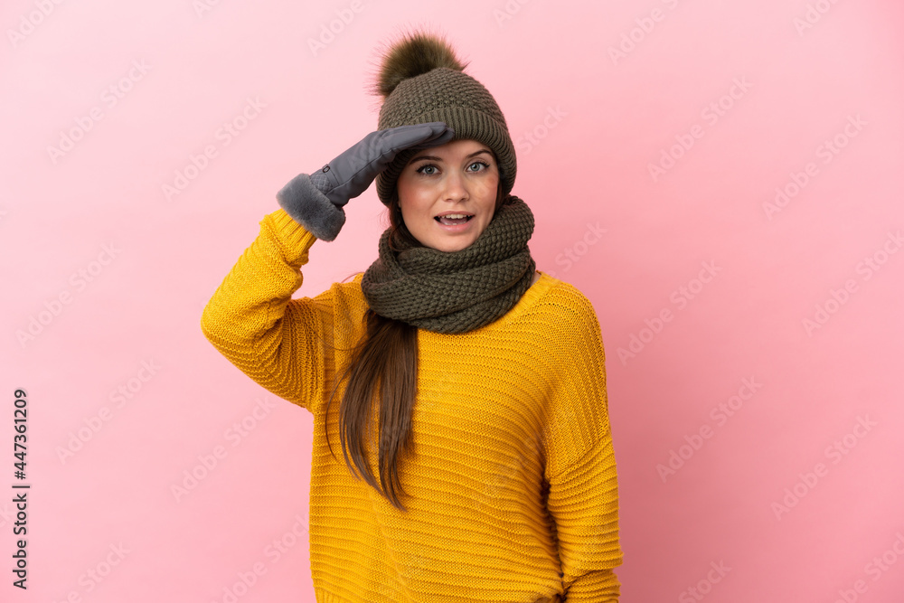 Young caucasian girl with winter hat isolated on pink background doing surprise gesture while looking to the side