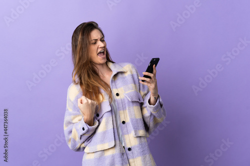 Young Ireland woman isolated on purple background using mobile phone and doing victory gesture