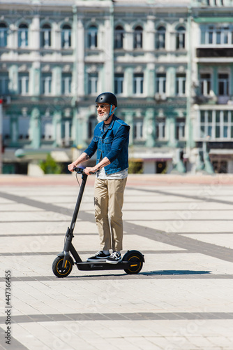 full length of cheerful man in sunglasses and helmet riding e-scooter in urban city