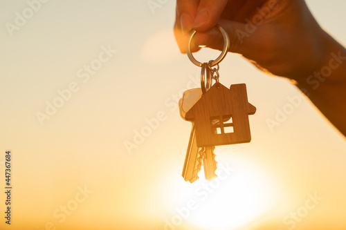 The hand holds the key to the new house with a keychain in the form of a house on the background of the sunset.