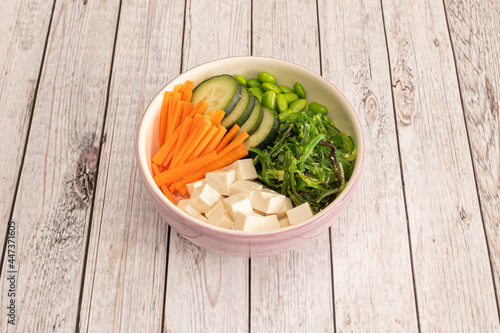 Pink vegan bowl with wakame seaweed salad with diced white tofu, grated carrot, cucumber slices and edamame beans on wooden table