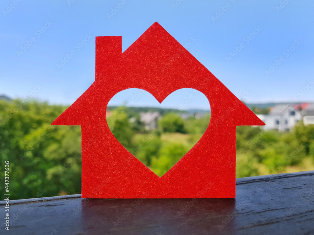 Small house cut out of red felt is standing on balcony railing. Heart-shaped hole is cut instead of a window. Blurred background of cottage village in summer sunny day. Dream home concept.