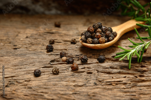 Black pepper in a wooden spoon with branch rosemary on a weathered wooden floor as background.