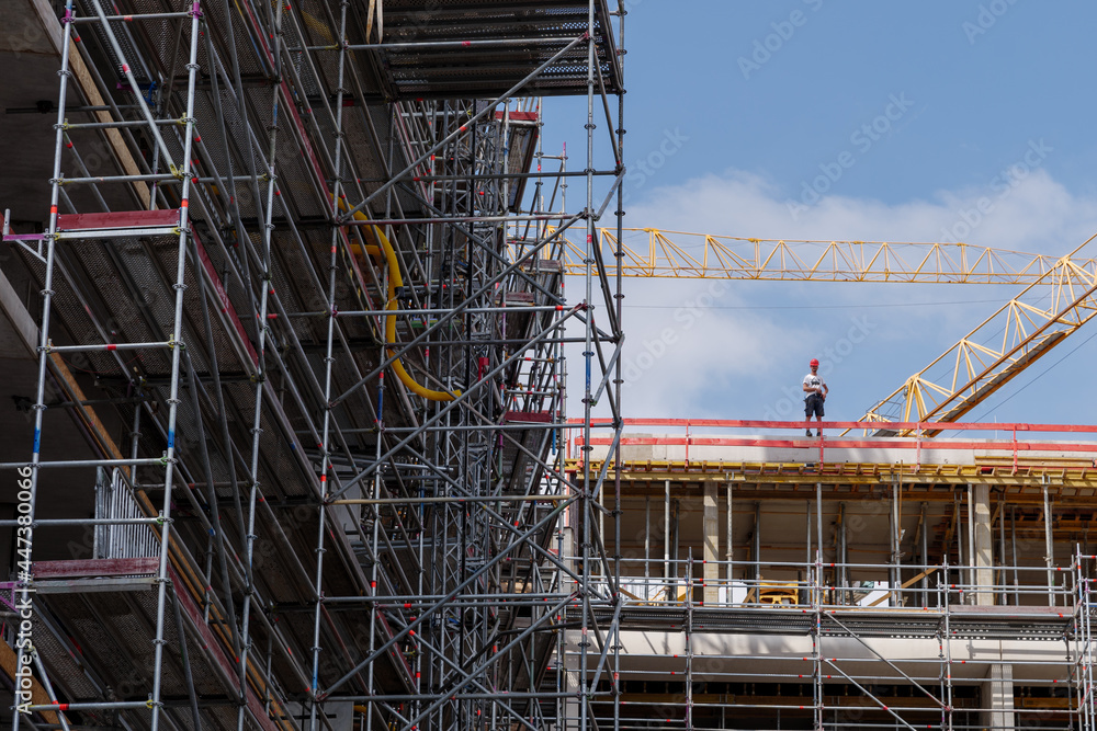Outdoor sunny view of construction building site covered with scaffold, people stand on building and crane against blue sky. 