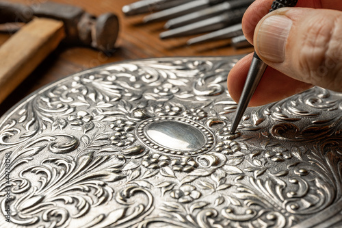 CLOSEUP OF CRAFTSMAN'S HAND EMBOSSING SILVER OR METAL WITH PUNCH IN THE WORKSHOP. GOLDSMITH, SILVERSMITH, JEWELLERY AND HANDICRAFTS CONCEPT. photo