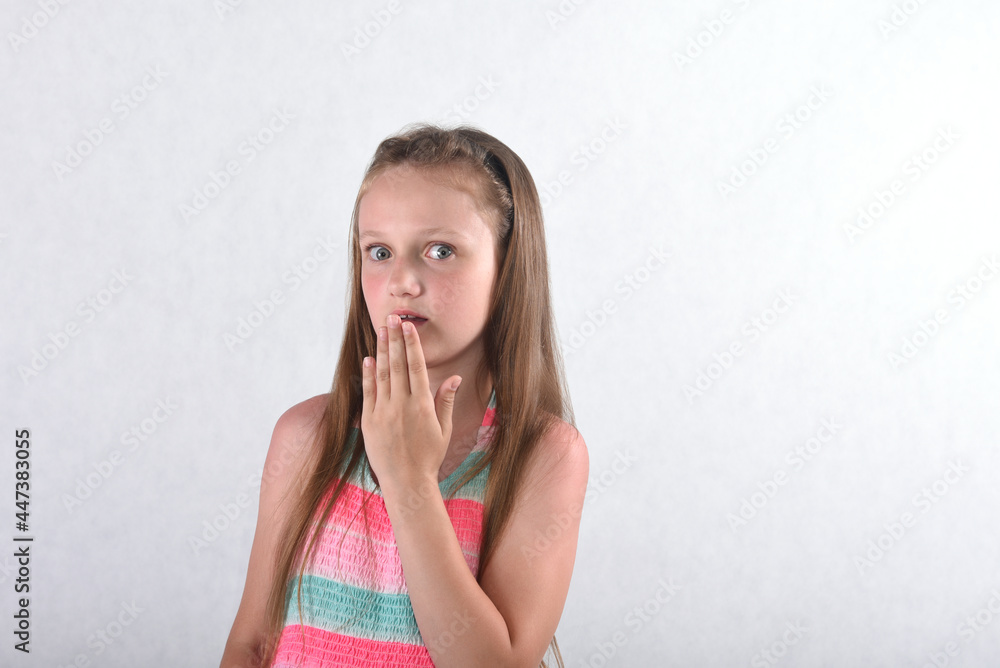 A beautiful school-age girl in the studio on a white background with a humorous face shows an emotion of surprise covering her mouth with her palm A beautiful school-age girl in the studio on a white background with a humorous face shows an emotion of surprise covering her mouth with her palm