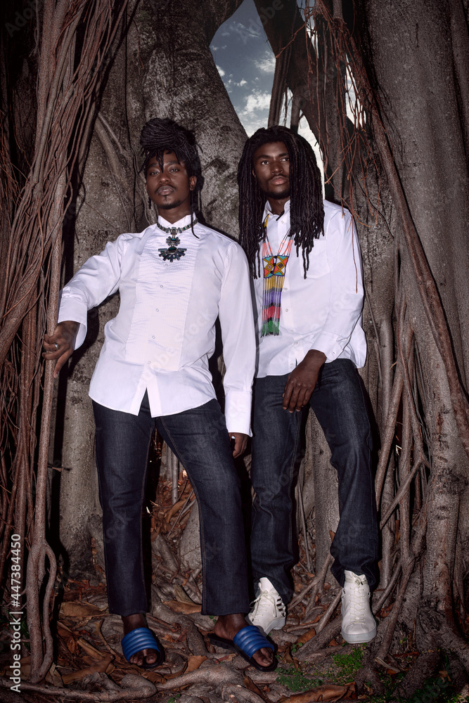 Portrait of two Millennial black man with dreadlocks posing by themselves outside leaning on an old oak tree wearing jeans, long sleeve shirts, gorgeous jewelry with blue sandals and white sneakers. Portrait of two Millennial black man with dreadlocks posing by themselves outside leaning on an old oak tree wearing jeans, long sleeve shirts, gorgeous jewelry with blue sandals and white sneakers.