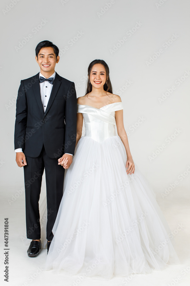 Full Length of young attractive Asian couple, soon to be bride and groom, woman wearing white wedding gown. Man wearing black tuxedo, standing together. Concept for pre wedding photography Full Length of young attractive Asian couple, soon to be bride and groom, woman wearing white wedding gown. Man wearing black tuxedo, standing together. Concept for pre wedding photography