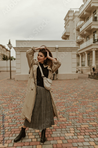 Tanned brunette woman touches hair and looks away outside. Full-length portrait of stylish lady in midi skirt and trench coat walks at street.