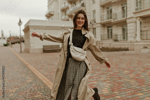 Tanned brunette woman in checkered skirt and black top smiles and moves outside. Charming happy girl in midi trench coat walks. photo