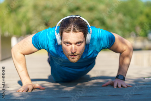 Front view of young strong athletic man wearing headphones and sportswear doing push ups on ground during sport training outdoors on hot summer day, focused male sportsman exercising in park
