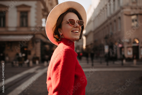Optimistic beautiful lady in beige hat and eyeglasses smiles outside. Charming woman in red sweater walks on street.